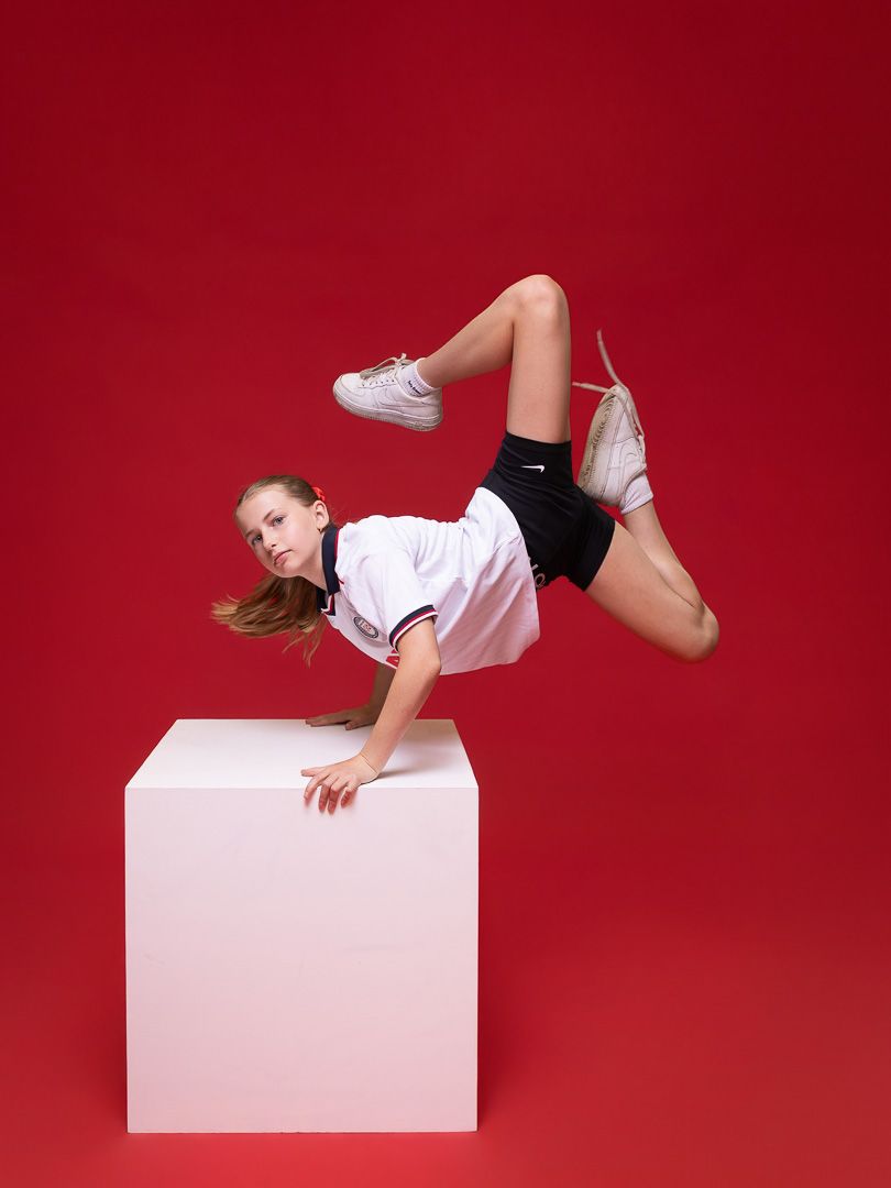A young girl in white shirt and black shorts performs a dynamic breakdance freeze pose on a white cube against red background.
