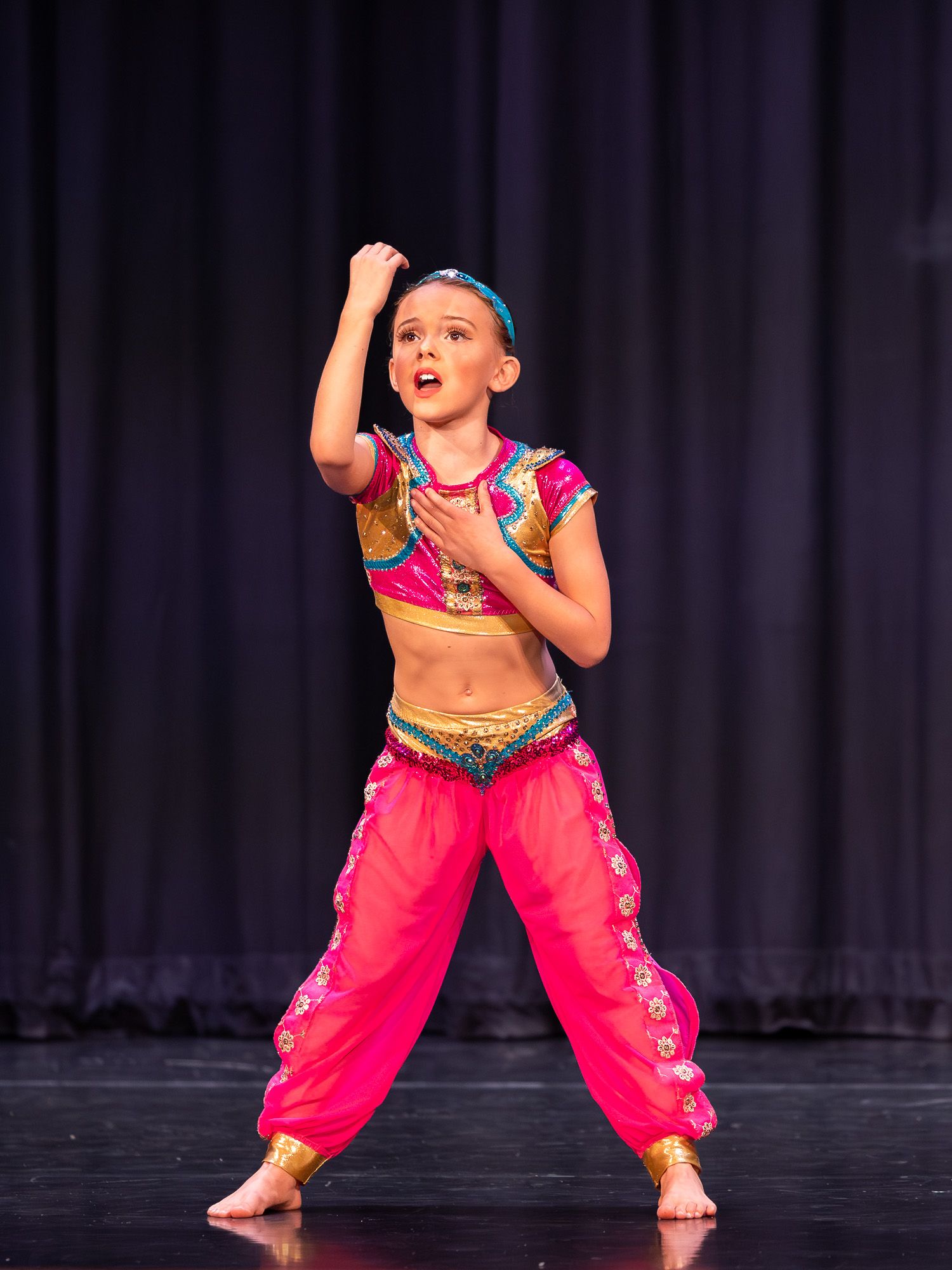 Young girl performs Indian classical dance on stage in vibrant pink and gold costume with expressive hand gestures.