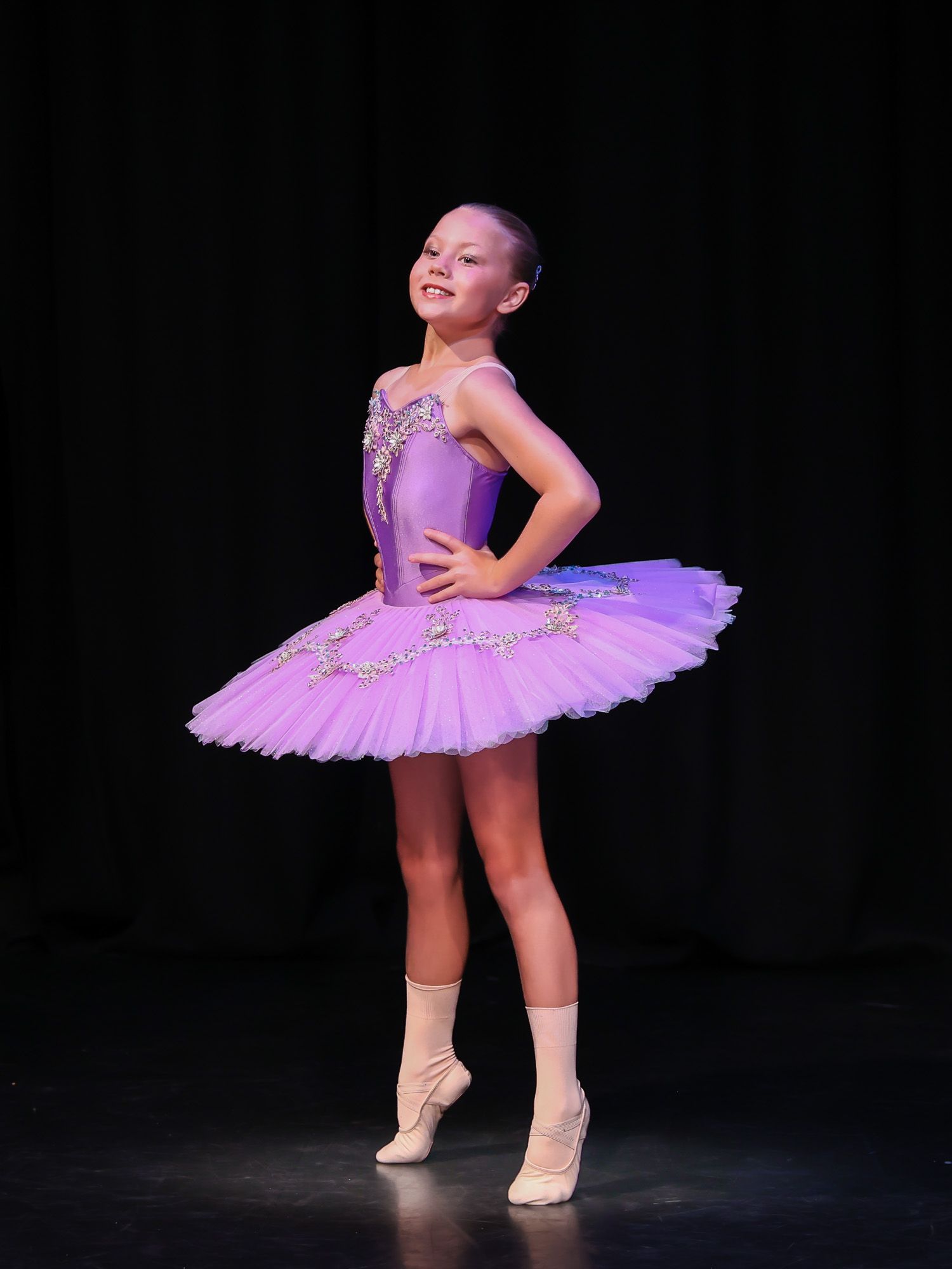 Young ballet dancer in purple tutu and pointe shoes poses gracefully on stage under spotlight.