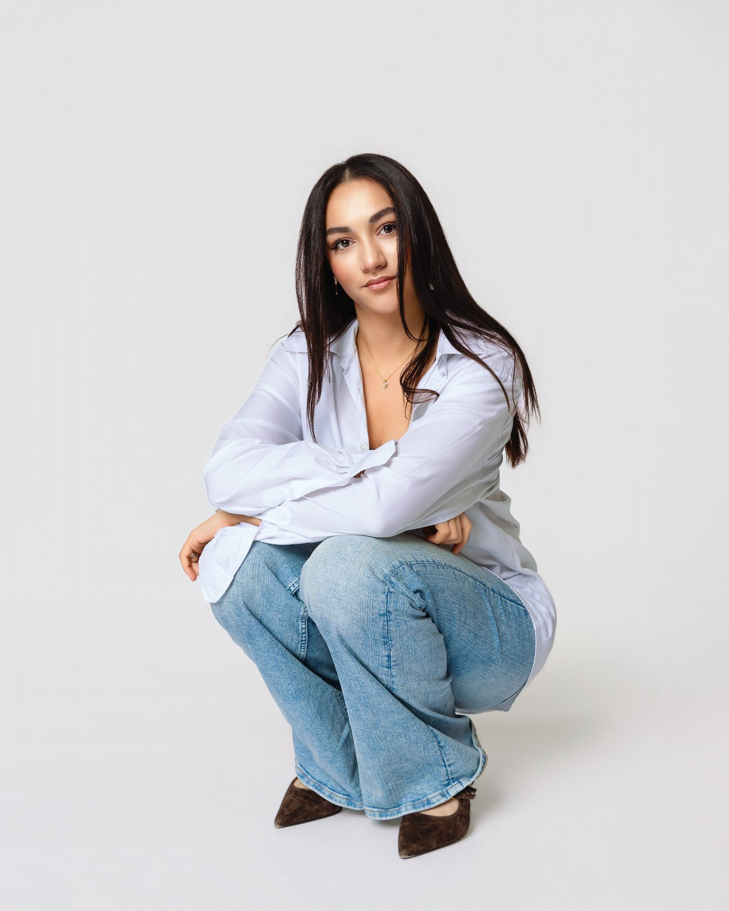 Person in white button-down shirt and baggy jeans crouches in casual pose against light background.