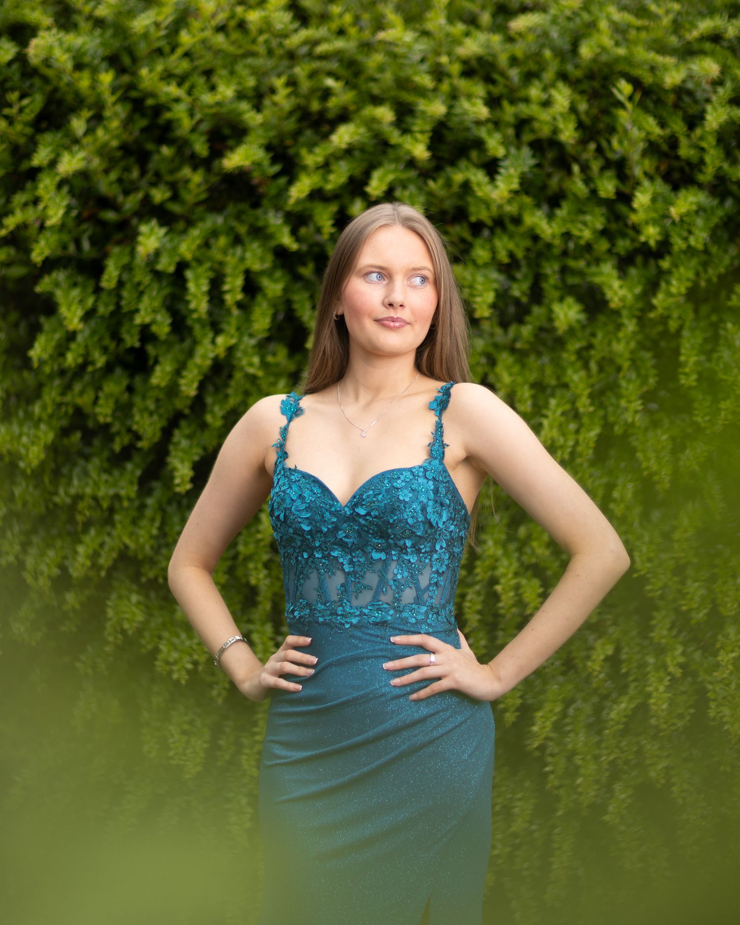 Elegant teal evening gown with lace bodice details photographed against lush green foliage.