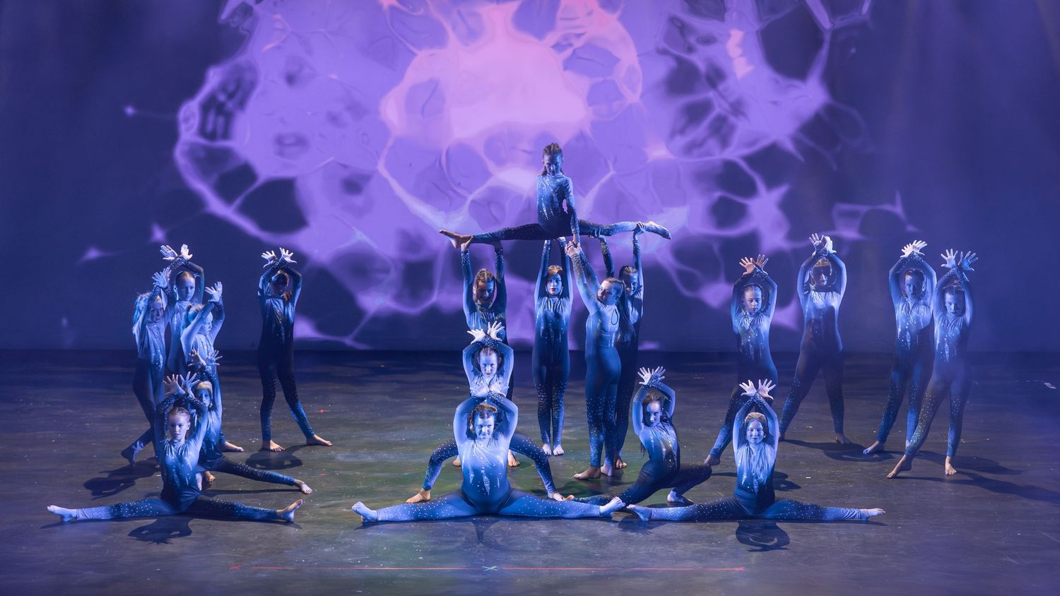 Dancers in blue costumes perform an artistic formation under purple lighting on a dark stage.