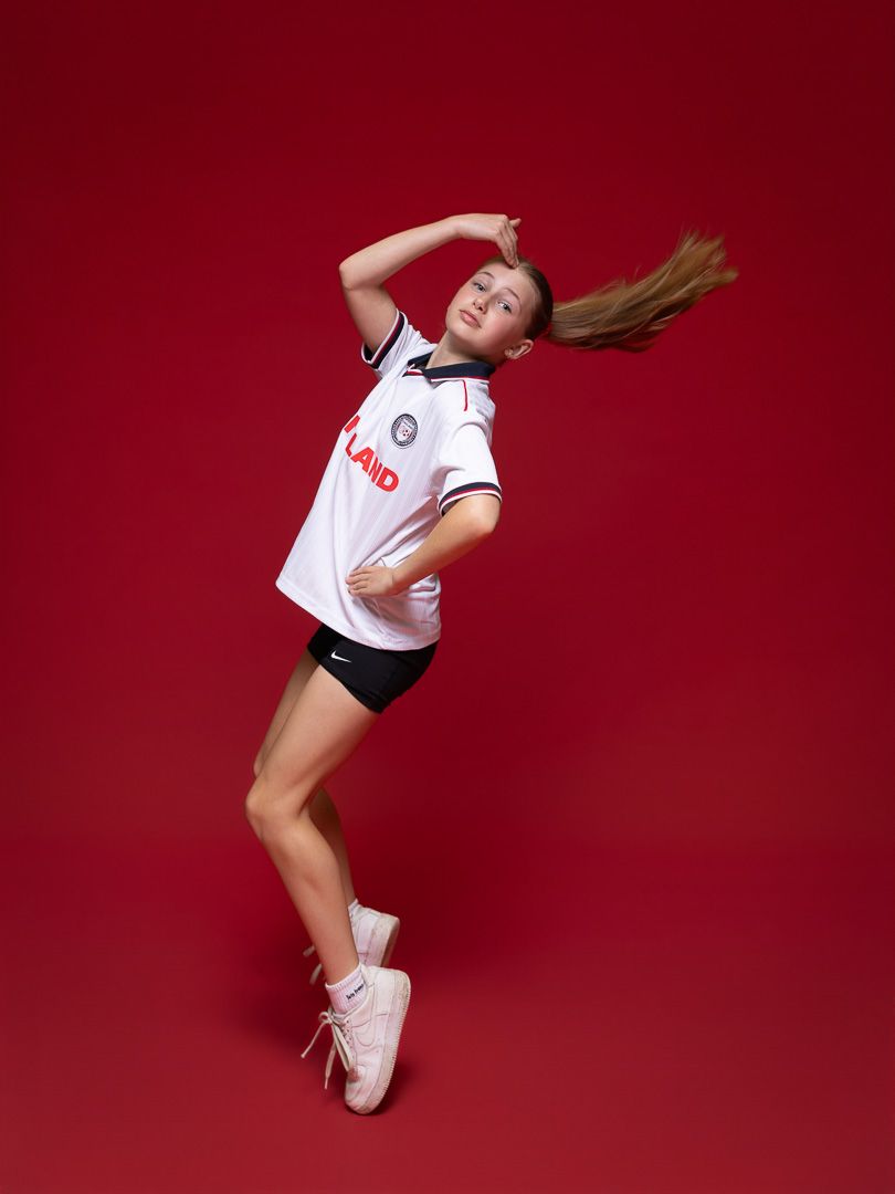 Athletic girl jumping in white soccer jersey and black shorts on red background.