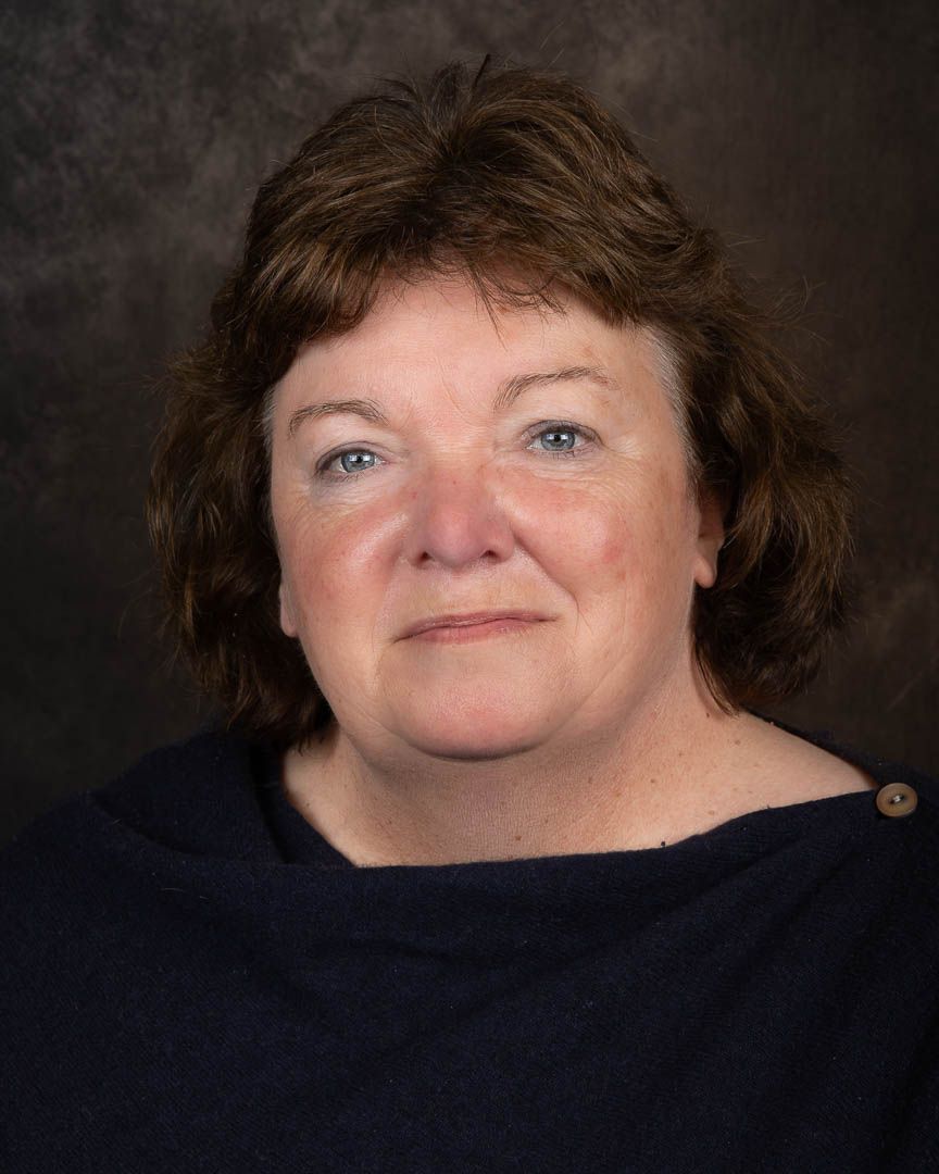 Professional headshot of a middle-aged woman with short brown hair wearing a navy top against a dark background.