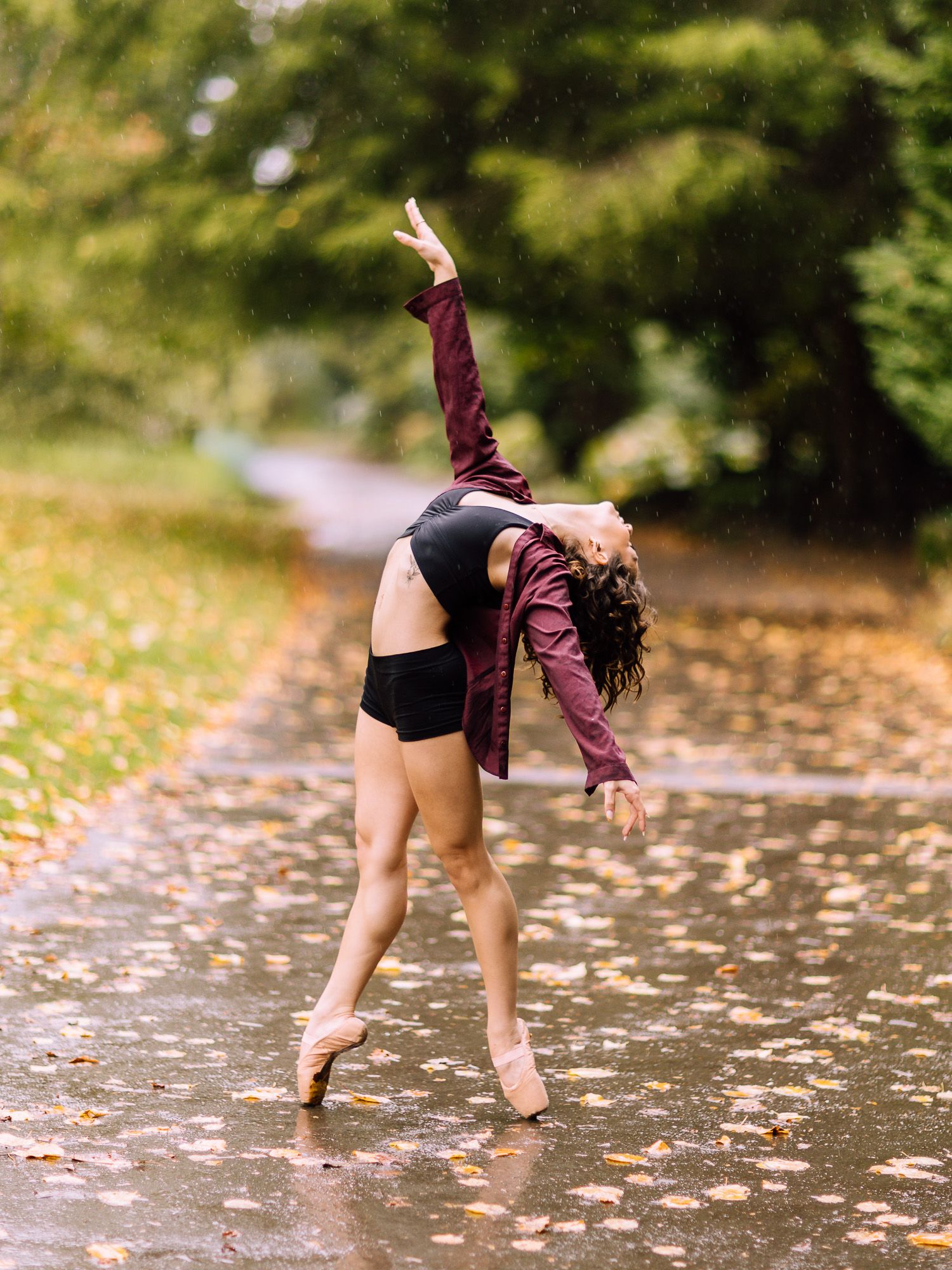 A ballet dancer in pointe shoes performs a graceful backbend on a rain-soaked path surrounded by autumn foliage.