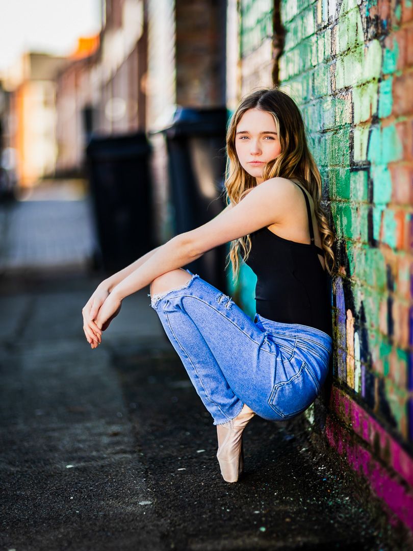 Dancer in denim and black top poses against colorful graffiti wall.