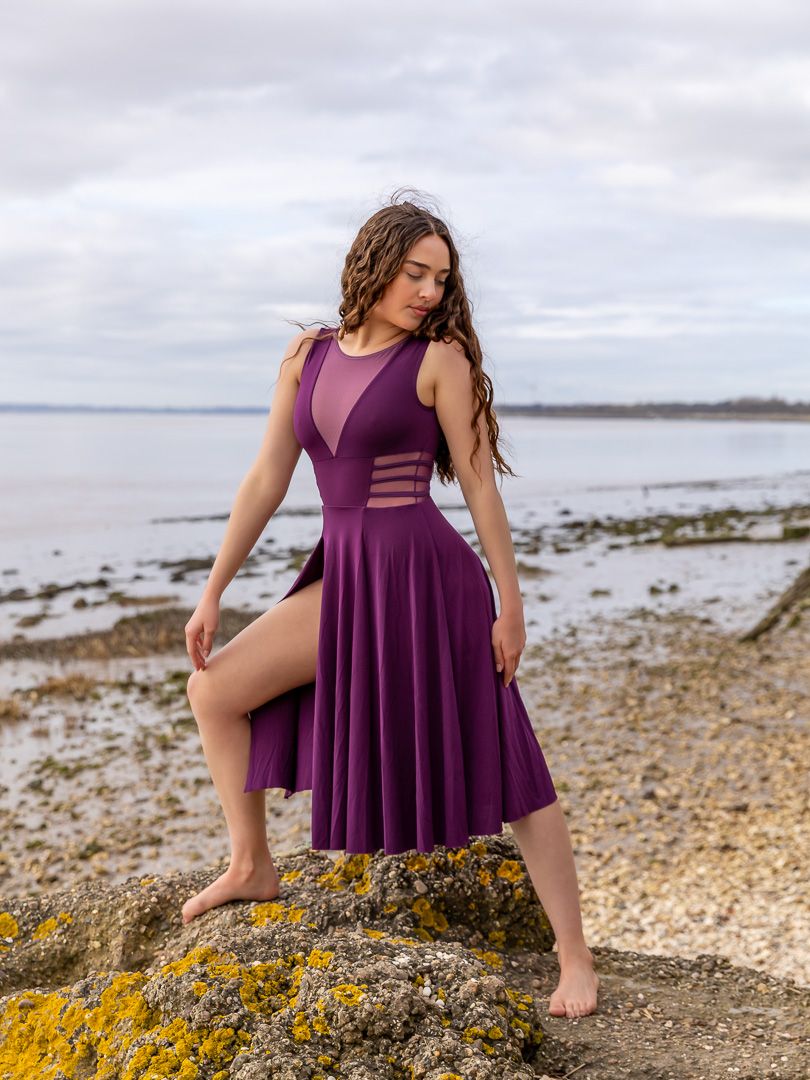 Dancer in purple dress poses expressively on rocky beach shoreline at sunset.