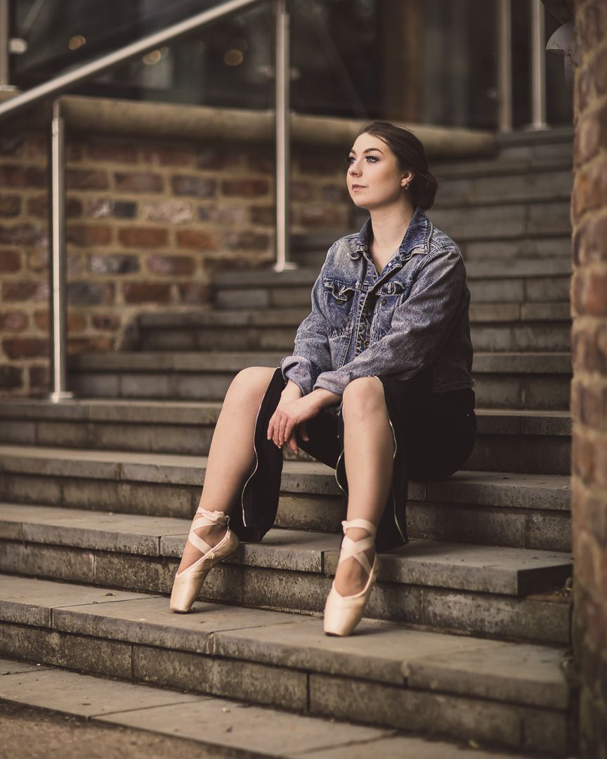 Ballet dancer sitting on stone steps wearing pointe shoes and denim jacket.