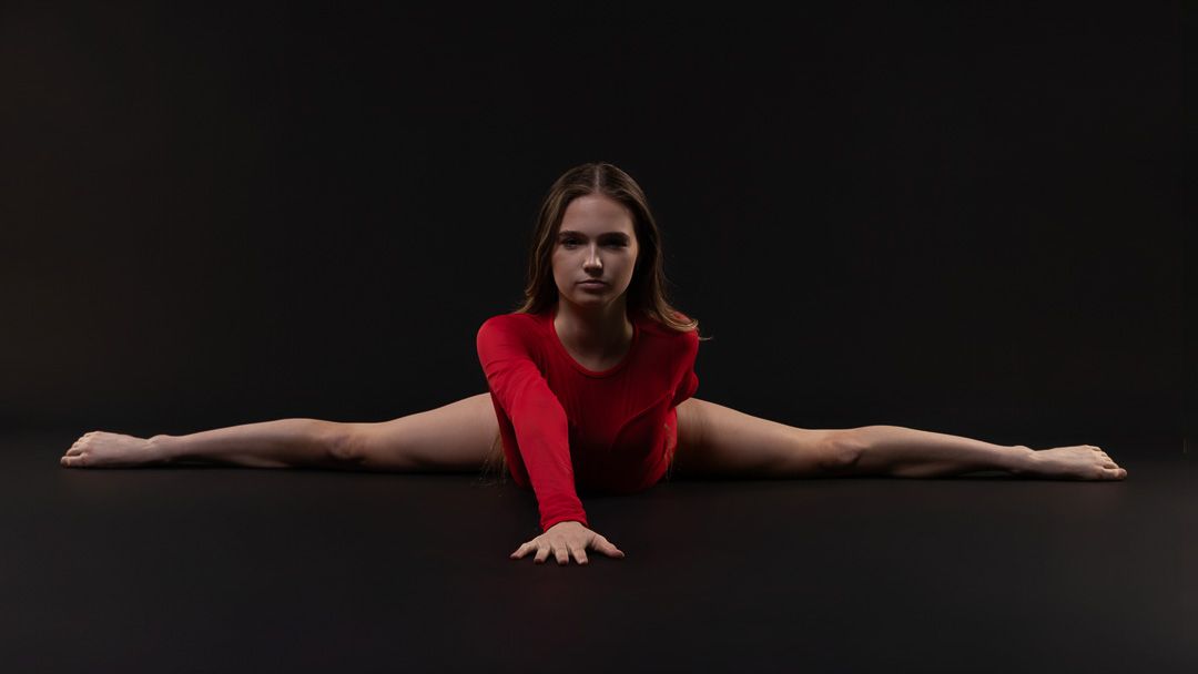 Flexible gymnast in red outfit performing a split on dark studio floor.