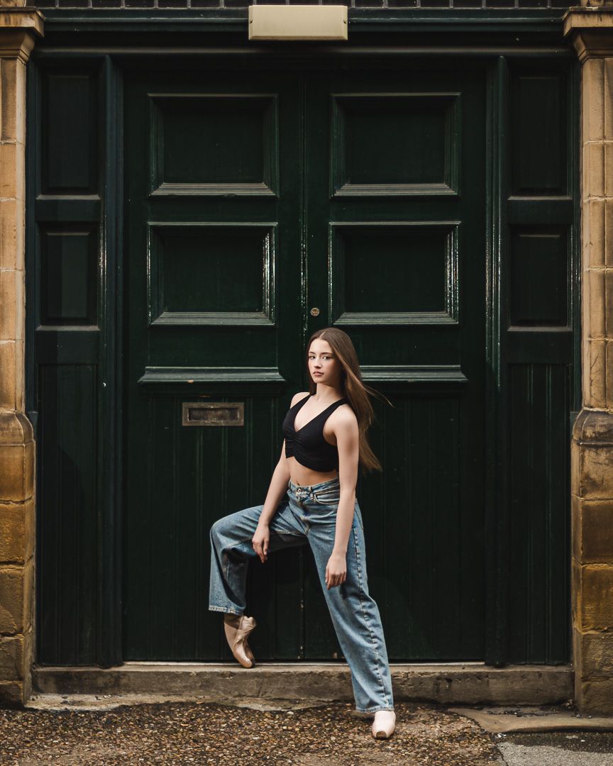 Young woman in black crop top and jeans poses gracefully on one foot before large dark green double doors.