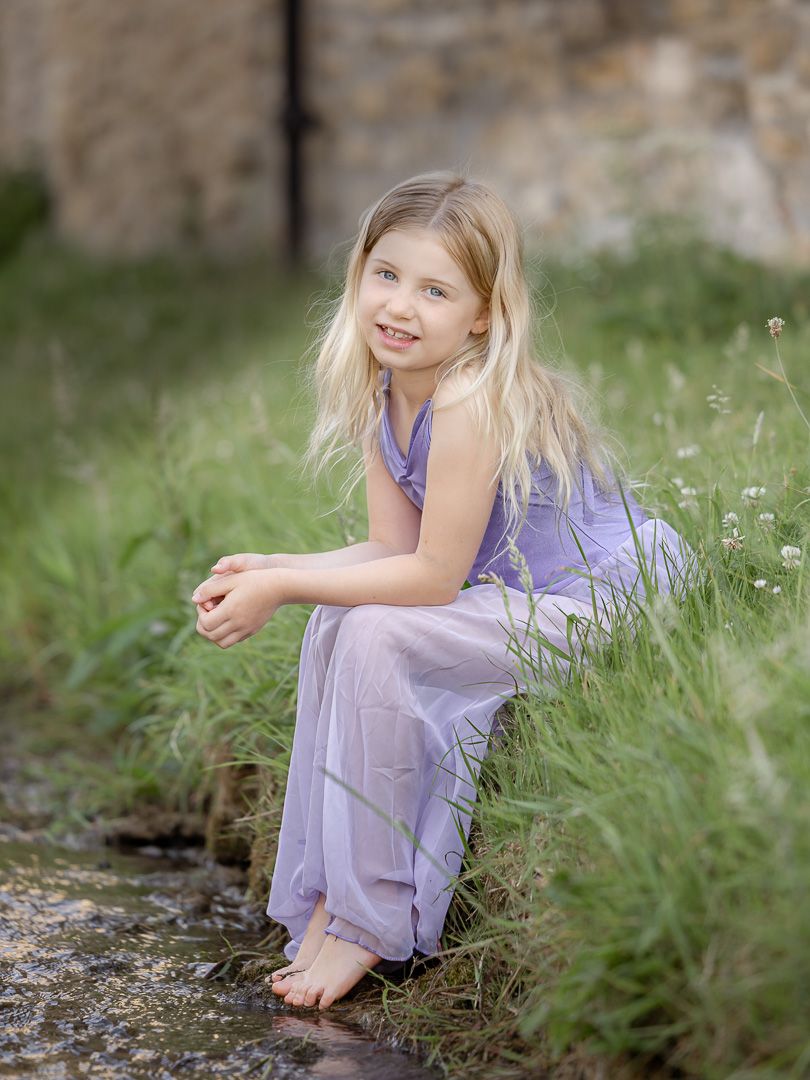 A child in a purple dress sits thoughtfully among wildflowers in a dreamy outdoor setting.