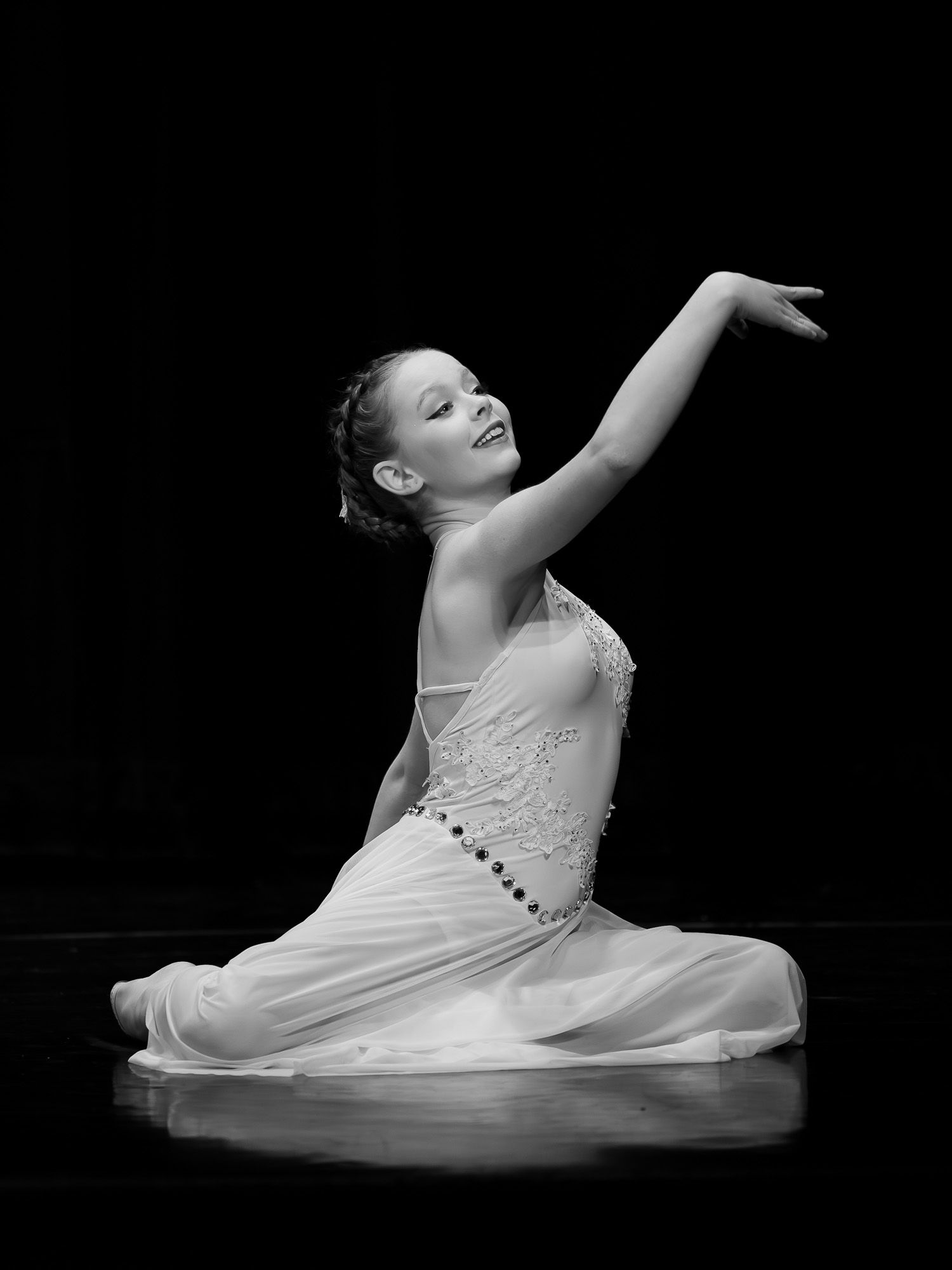 Young ballet dancer in white dress performs graceful floor pose with arm raised, captured in black and white photography.