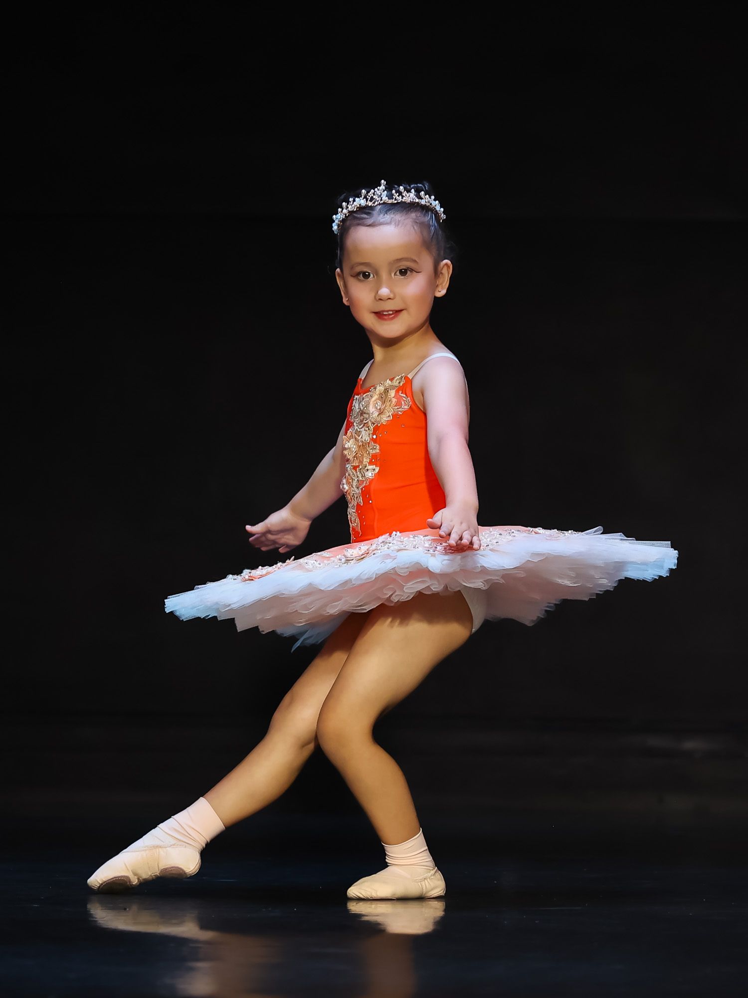 Young girl in orange ballet tutu and tiara performing on stage in ballet recital.