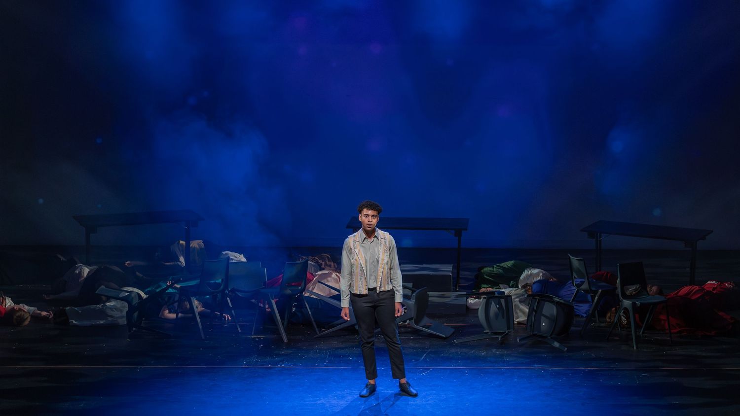 A lone performer stands center stage under blue dramatic lighting in a theatrical production set.