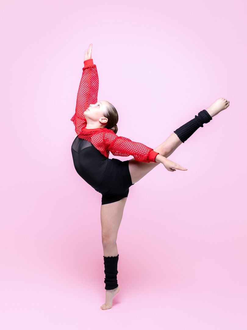 Young dancer in red mesh top and black shorts performs a high kick against a pink background.