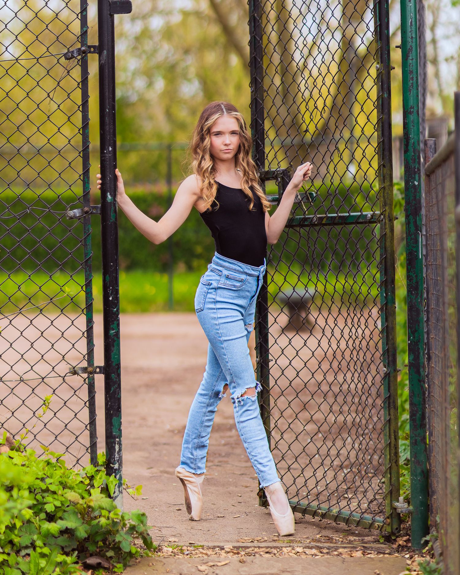 A person in jeans poses gracefully against a chain link fence in a park setting during spring.