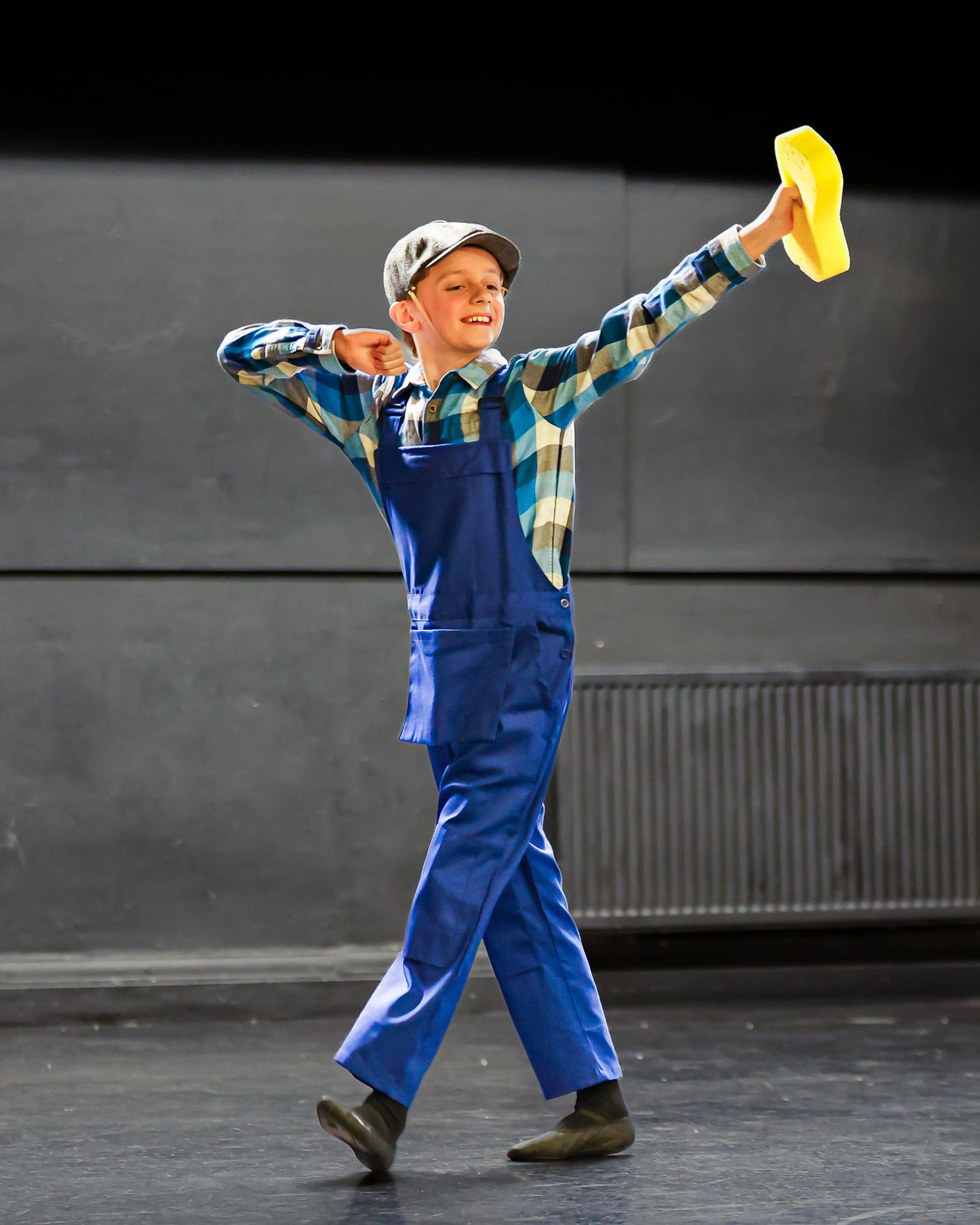 Boy in blue overalls and plaid shirt dancing on stage, holding a yellow hat with arm raised.