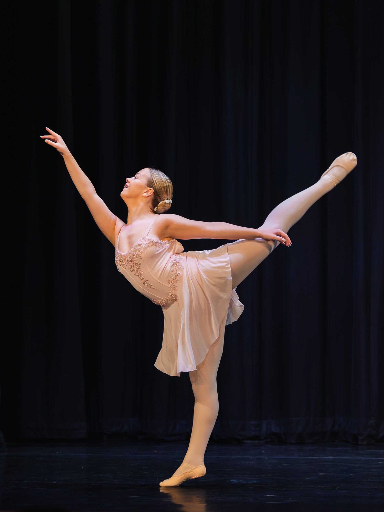 A ballet dancer performs an arabesque on stage in a pink lyrical costume under professional stage lighting.