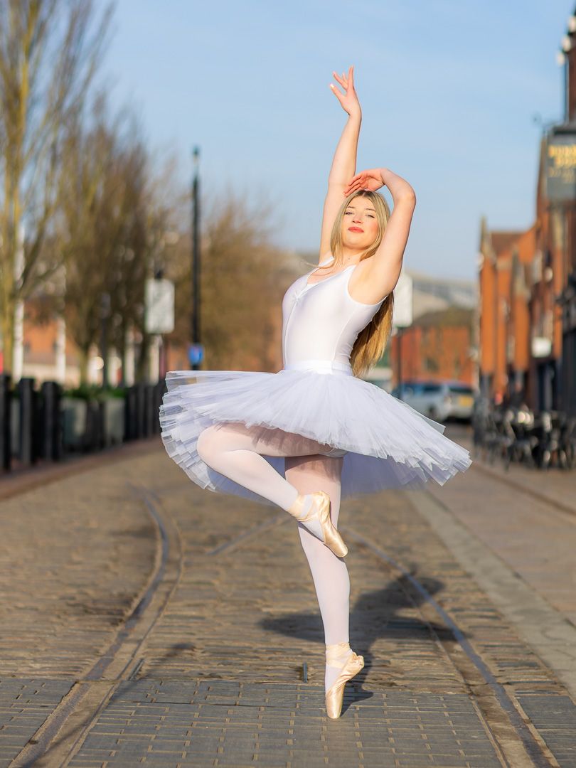 Dancer in white tutu performs ballet moves on cobblestone street.
