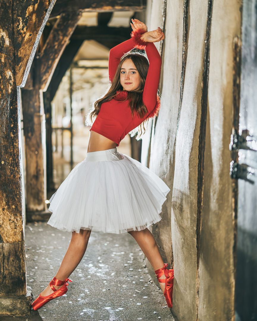 A ballet dancer in red pointe shoes and white tutu poses gracefully between wooden pier posts at sunset.