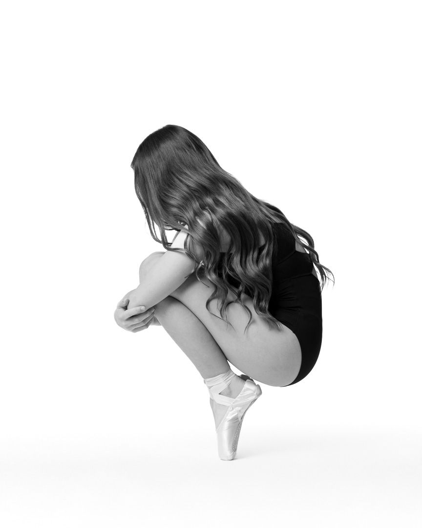 Black and white photo of a ballerina in pointe shoes, crouched with knees drawn up, long hair cascading forward.