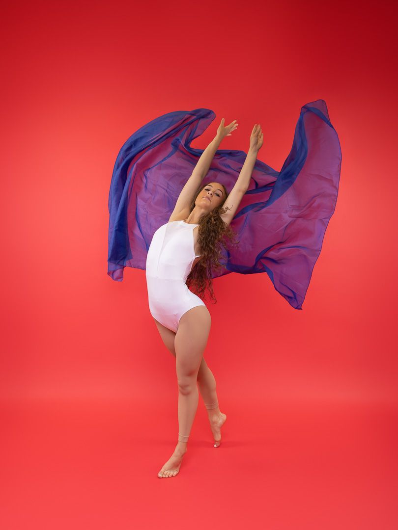 A dancer in a white leotard poses gracefully against a red background, holding a flowing blue and purple fabric overhead.