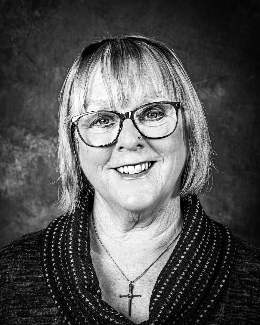 Black and white professional headshot of a smiling woman with bob haircut, glasses, and cross necklace.