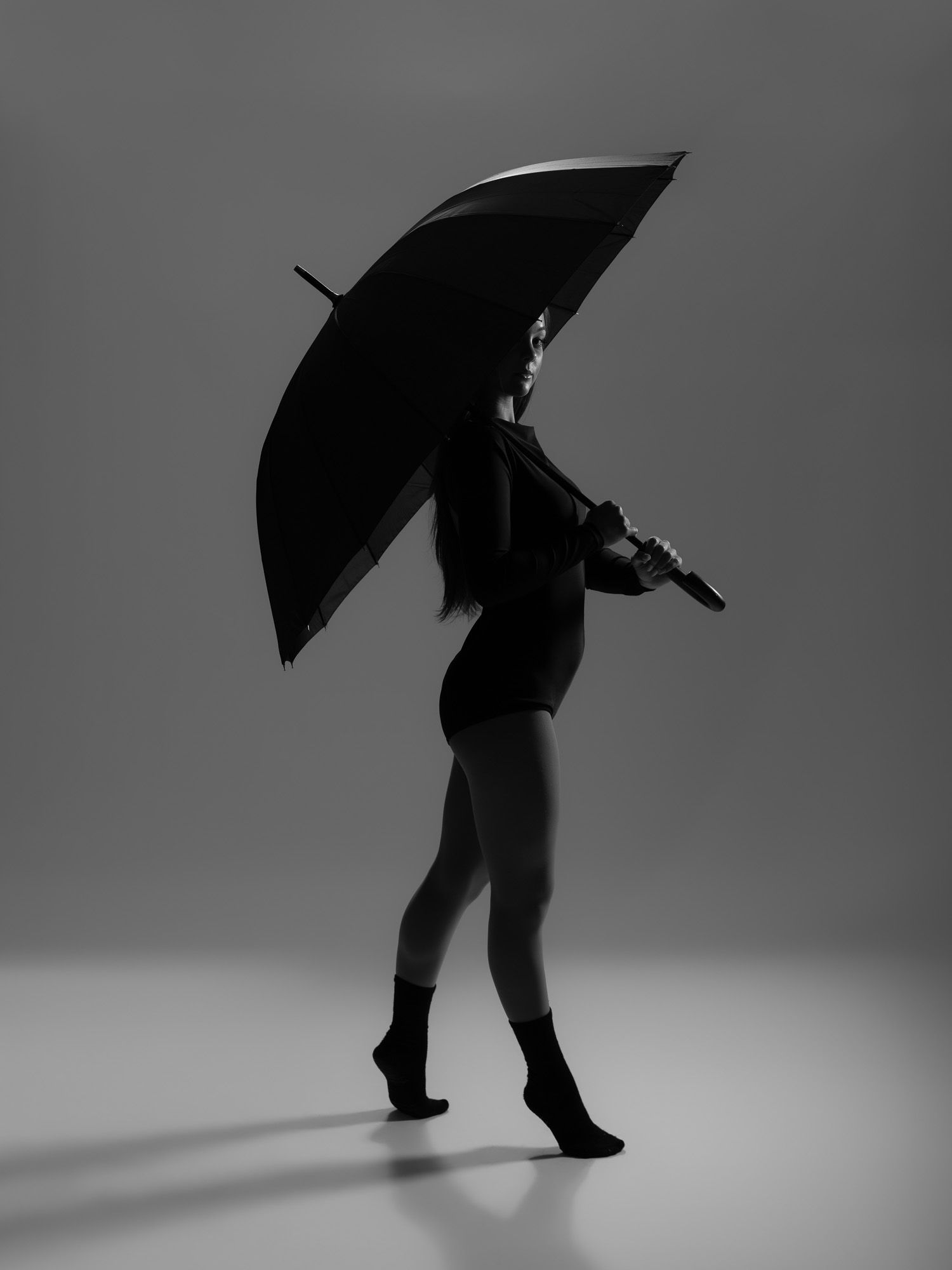 Black and white silhouette of a woman holding an open umbrella, wearing a bodysuit and socks, studio photography.