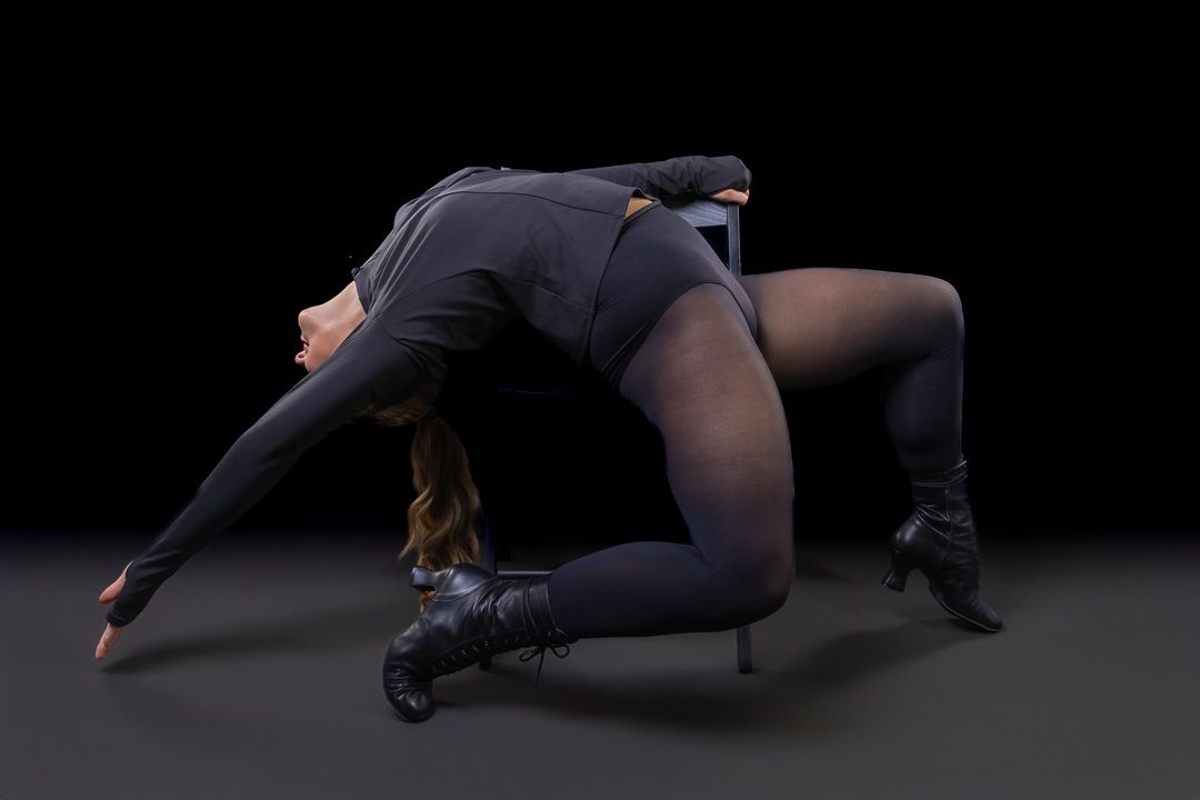 A flexible dancer in all-black attire performs a dramatic backbend on a dark studio floor.