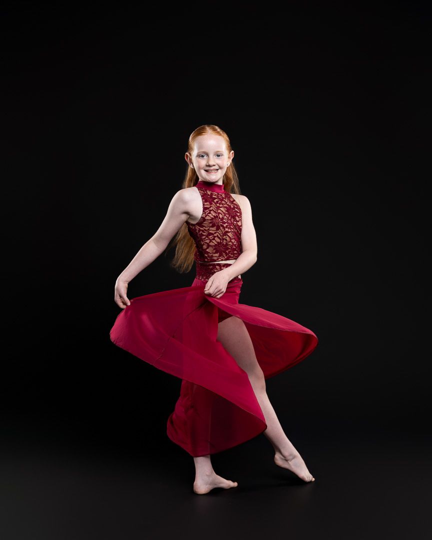 Young redhead girl dancing in elegant red lace gown against black background.