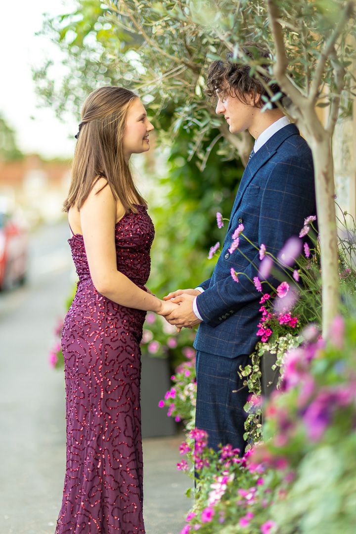 A couple in formal attire share a romantic moment among purple flowers and greenery.