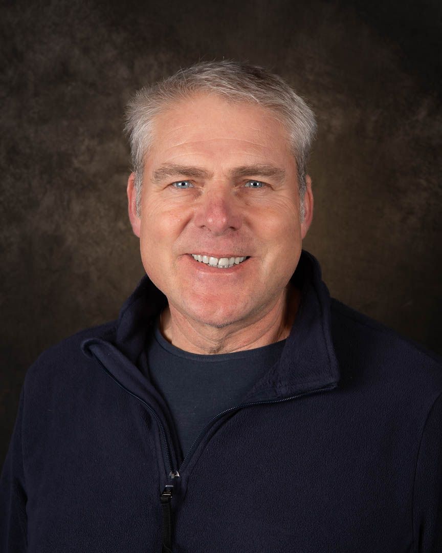 Smiling middle-aged man with gray hair wearing a navy blue fleece jacket, photographed against a dark studio background.