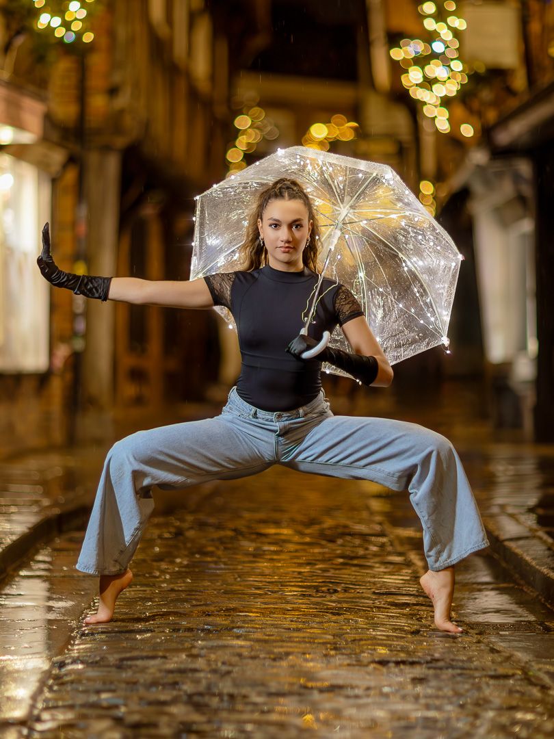 Dance performance with clear umbrella on wet cobblestone street with bokeh lights.