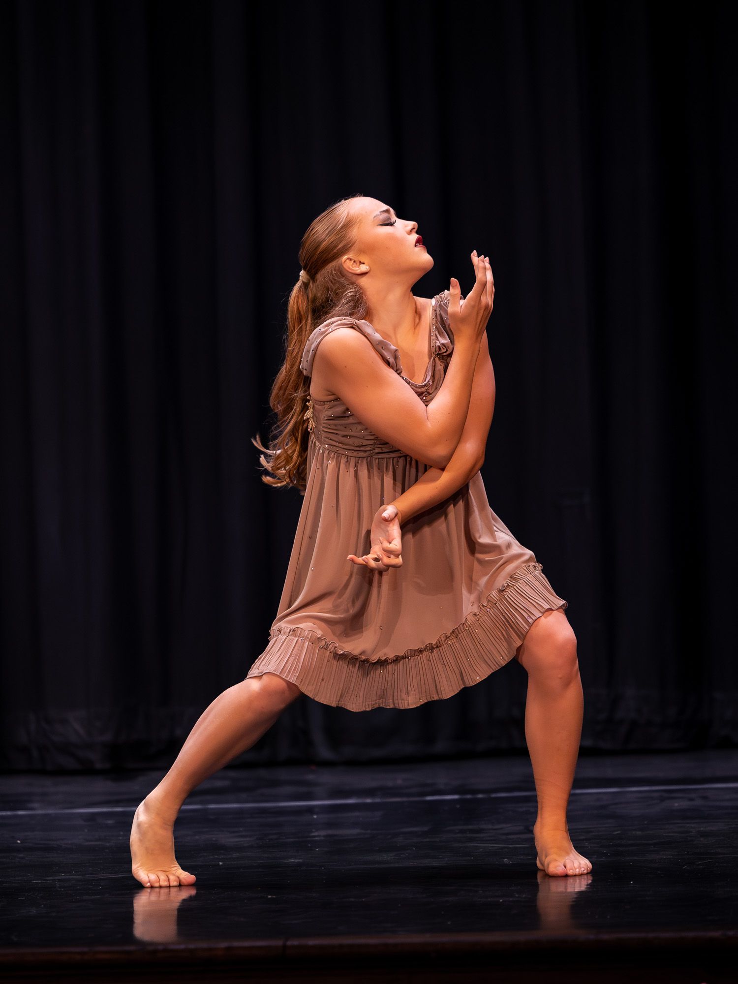 Young dancer performs expressive contemporary dance on stage, wearing a tan dress, barefoot under dramatic lighting.