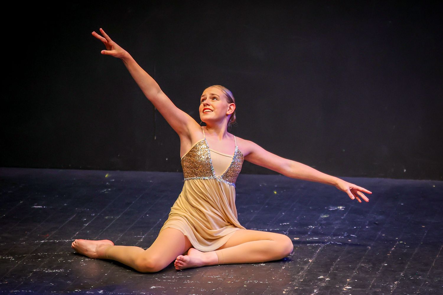 Young dancer in gold costume performs graceful floor routine on stage, arm extended upward under dramatic lighting.