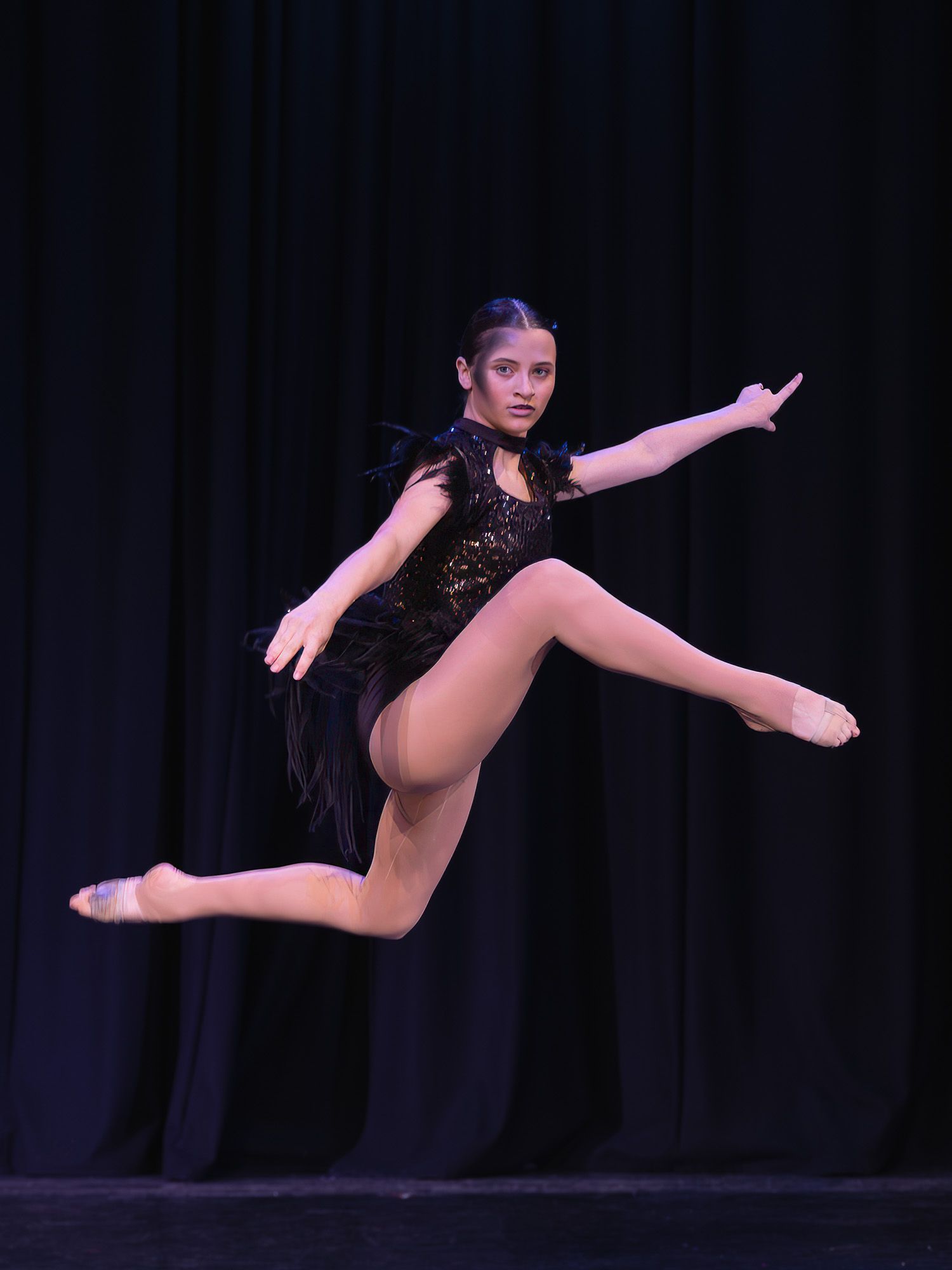 Dancer in black feathered costume leaps gracefully mid-air on stage during performance.