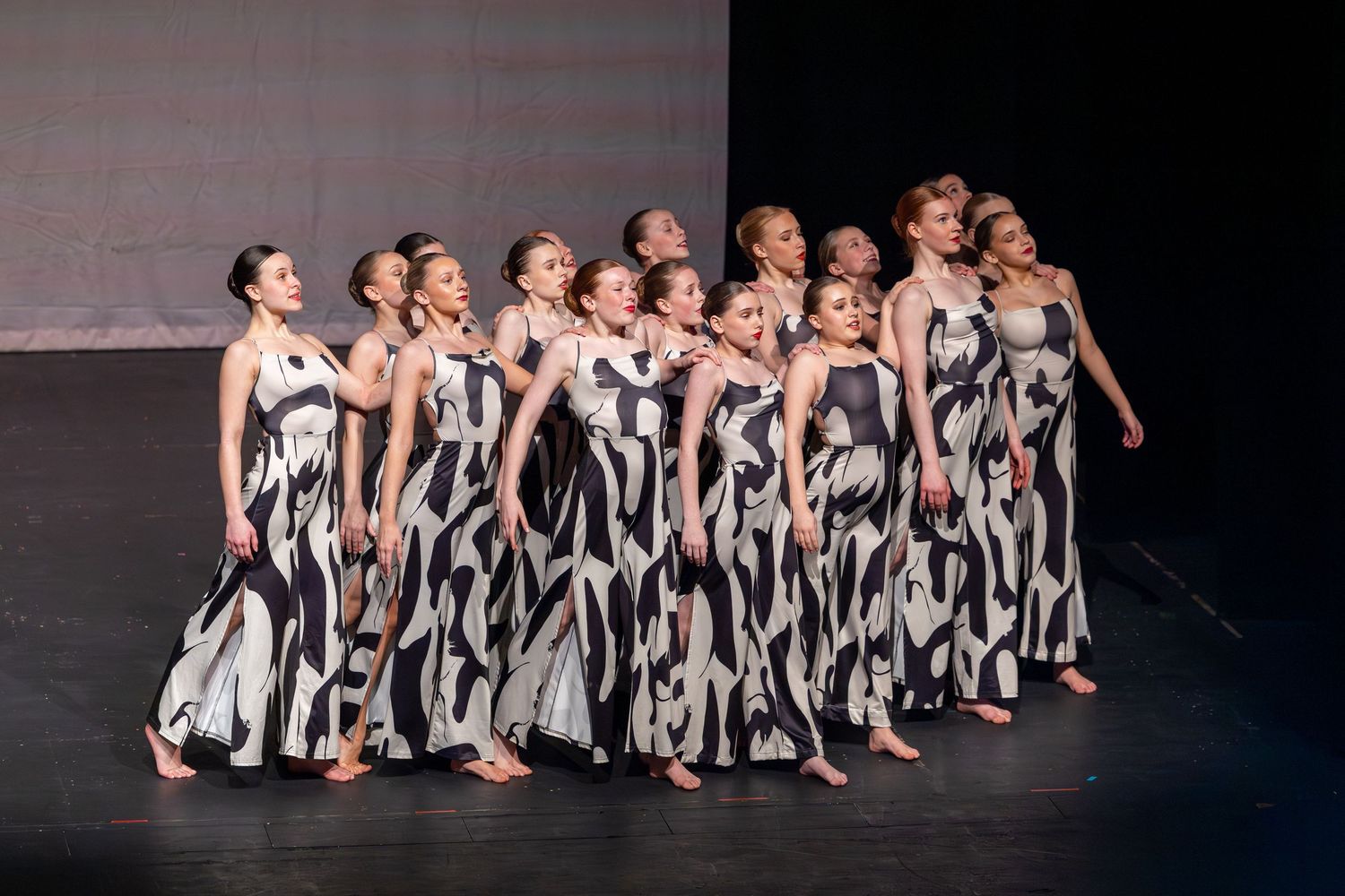 A group of dancers perform on stage wearing matching black and white patterned gowns under dramatic stage lighting.