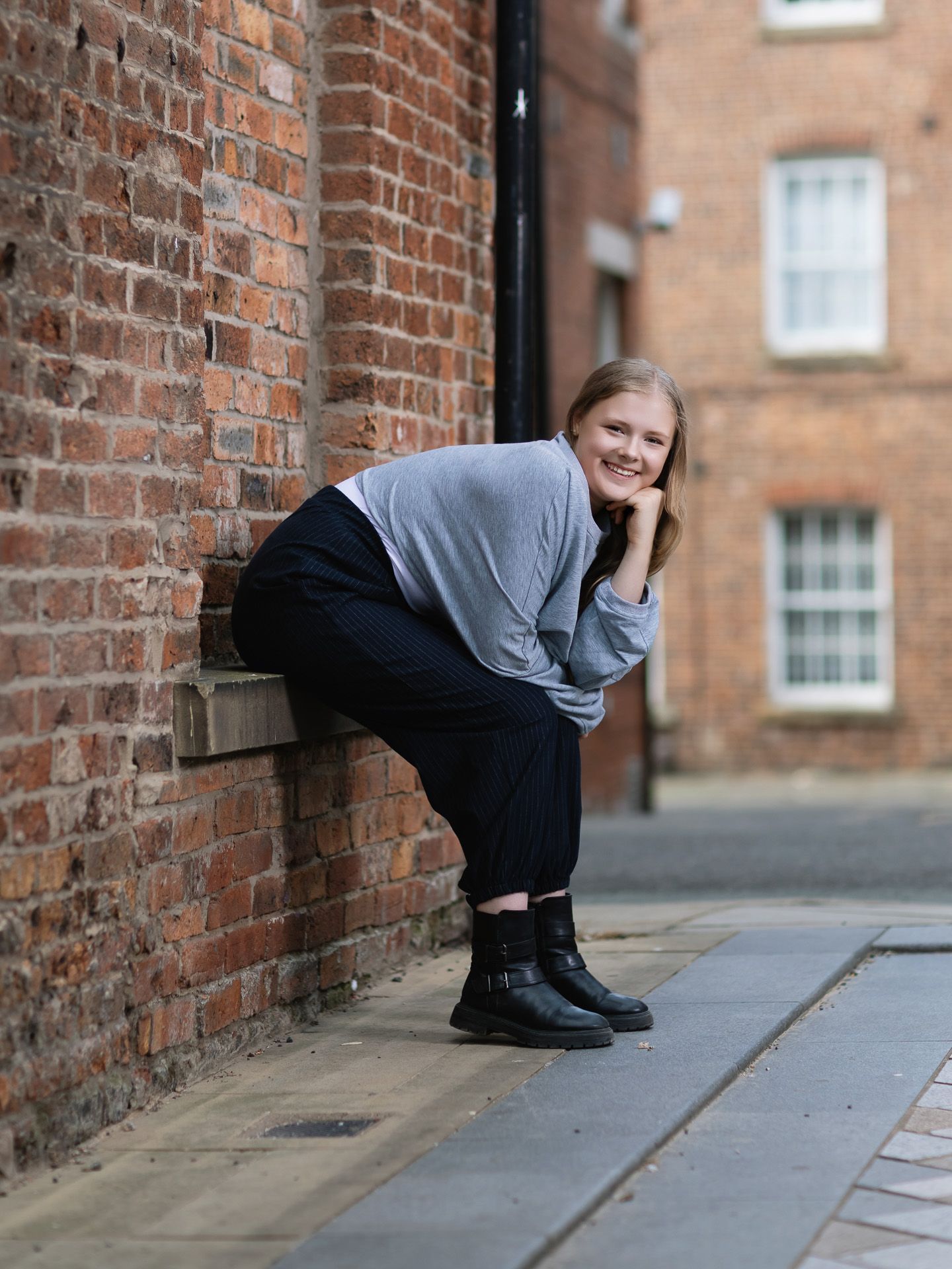 Young woman smiling and posing against a red brick wall on a cobblestone street wearing a grey sweater and black pants.