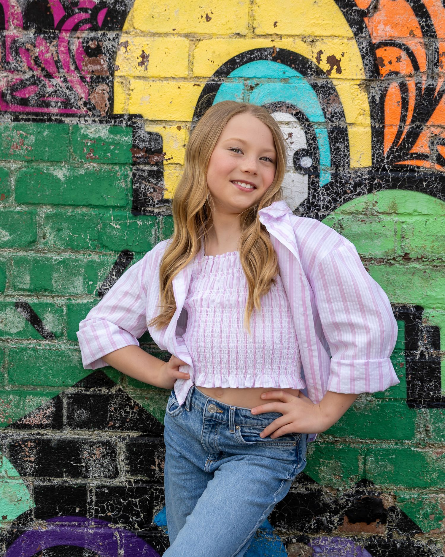 Young girl in pink top and jeans posing confidently in front of colorful graffiti brick wall.
