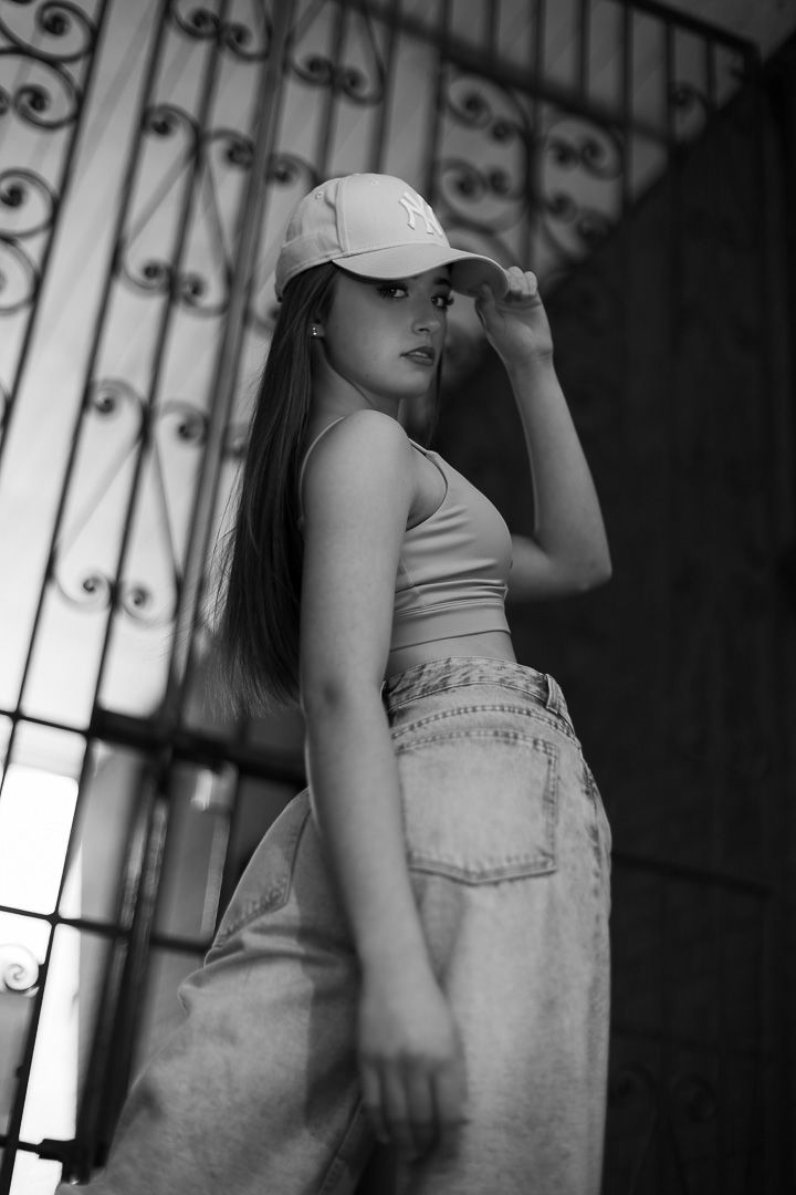 Black and white portrait of a young woman in cap, crop top, and baggy jeans posing by ornate metal gates.