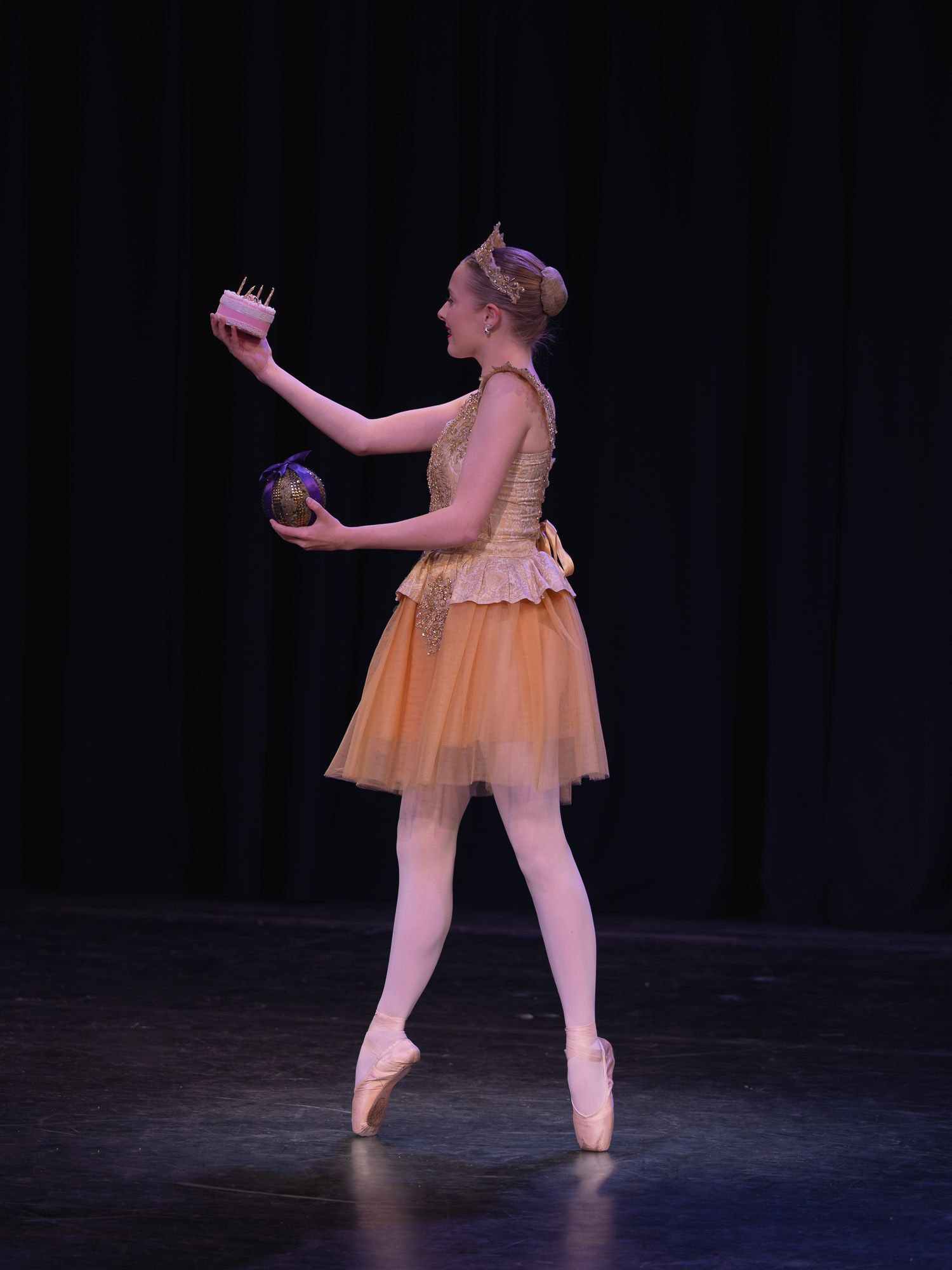 Ballet dancer en pointe in orange tutu holds props on stage under spotlight during performance.