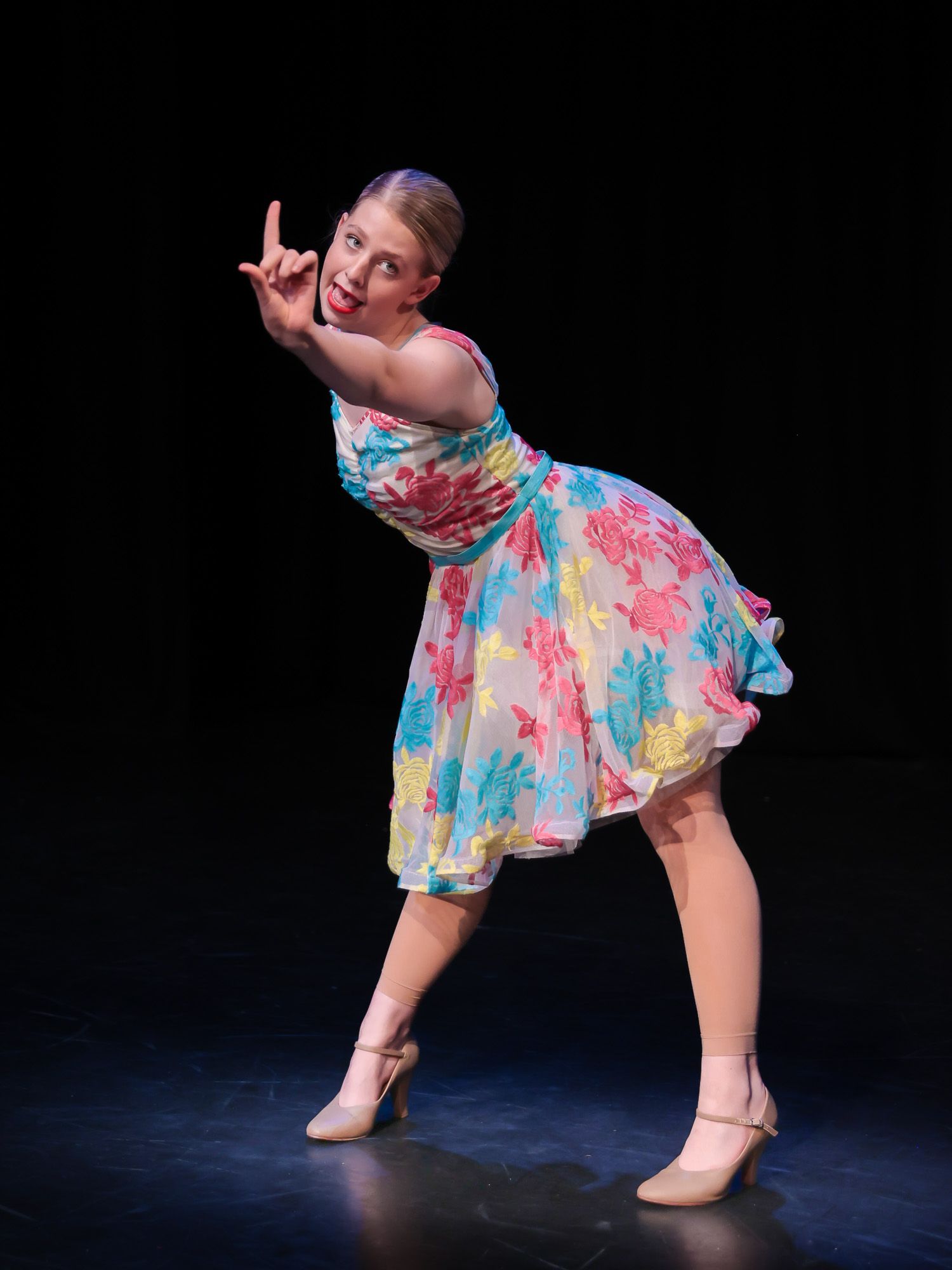 Young girl in floral dress performs expressive dance pose on stage under spotlight.