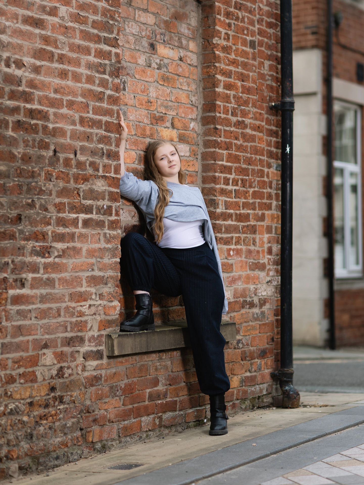 Young woman in grey top and black joggers posing against a red brick wall on a cobblestone street.