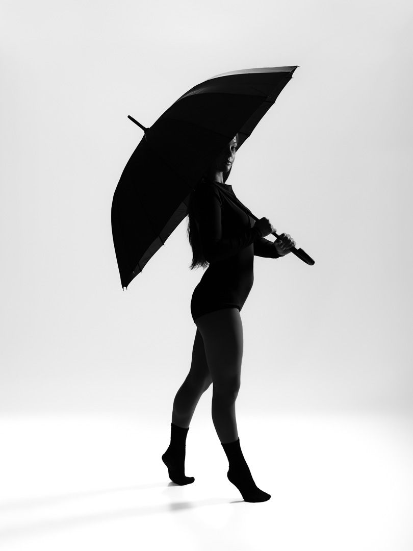 Silhouette of a woman holding a large black umbrella, posed gracefully against a white background in a black and white photo.