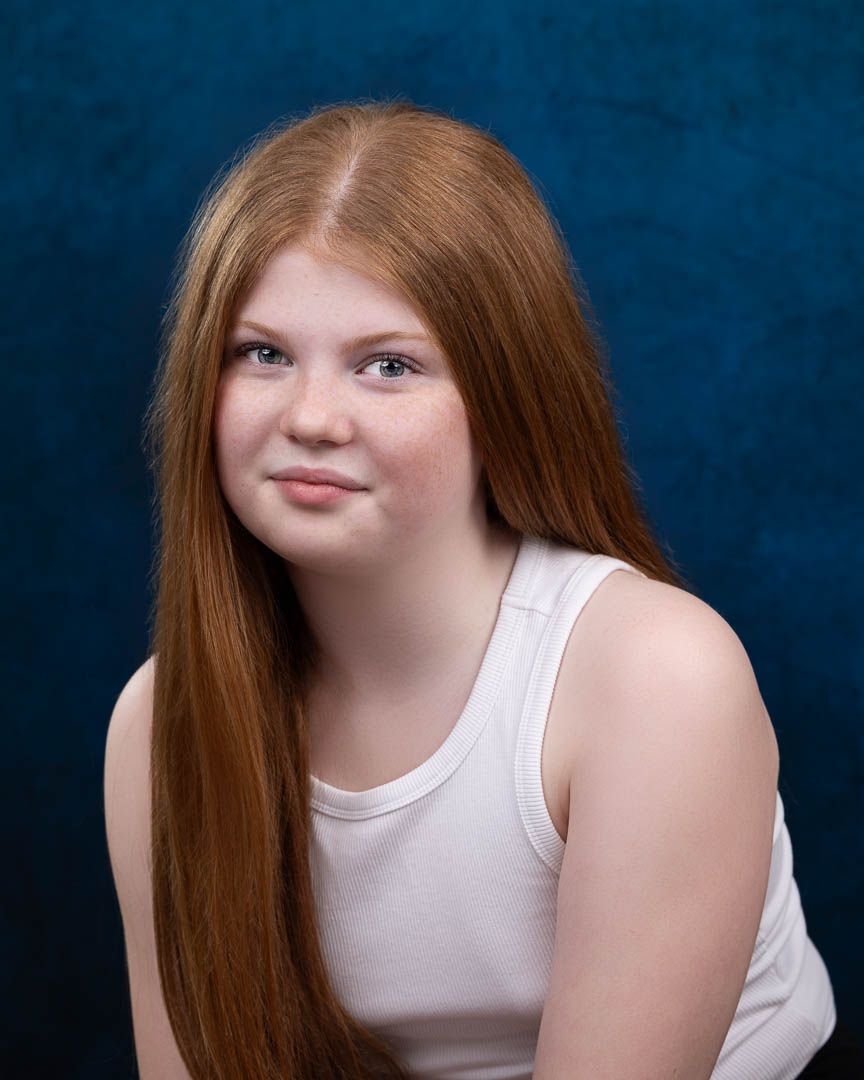 Young girl with long red hair smiling in a white tank top against a dark blue background.