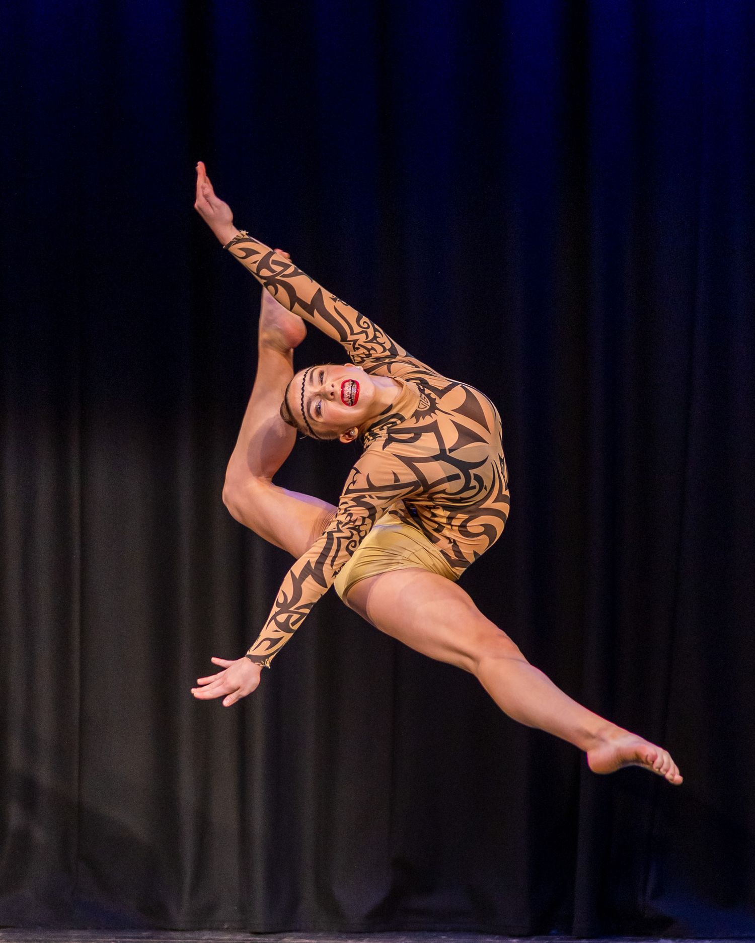 Dancer in gold and black patterned leotard performs a stunning aerial leap on stage with leg raised overhead.