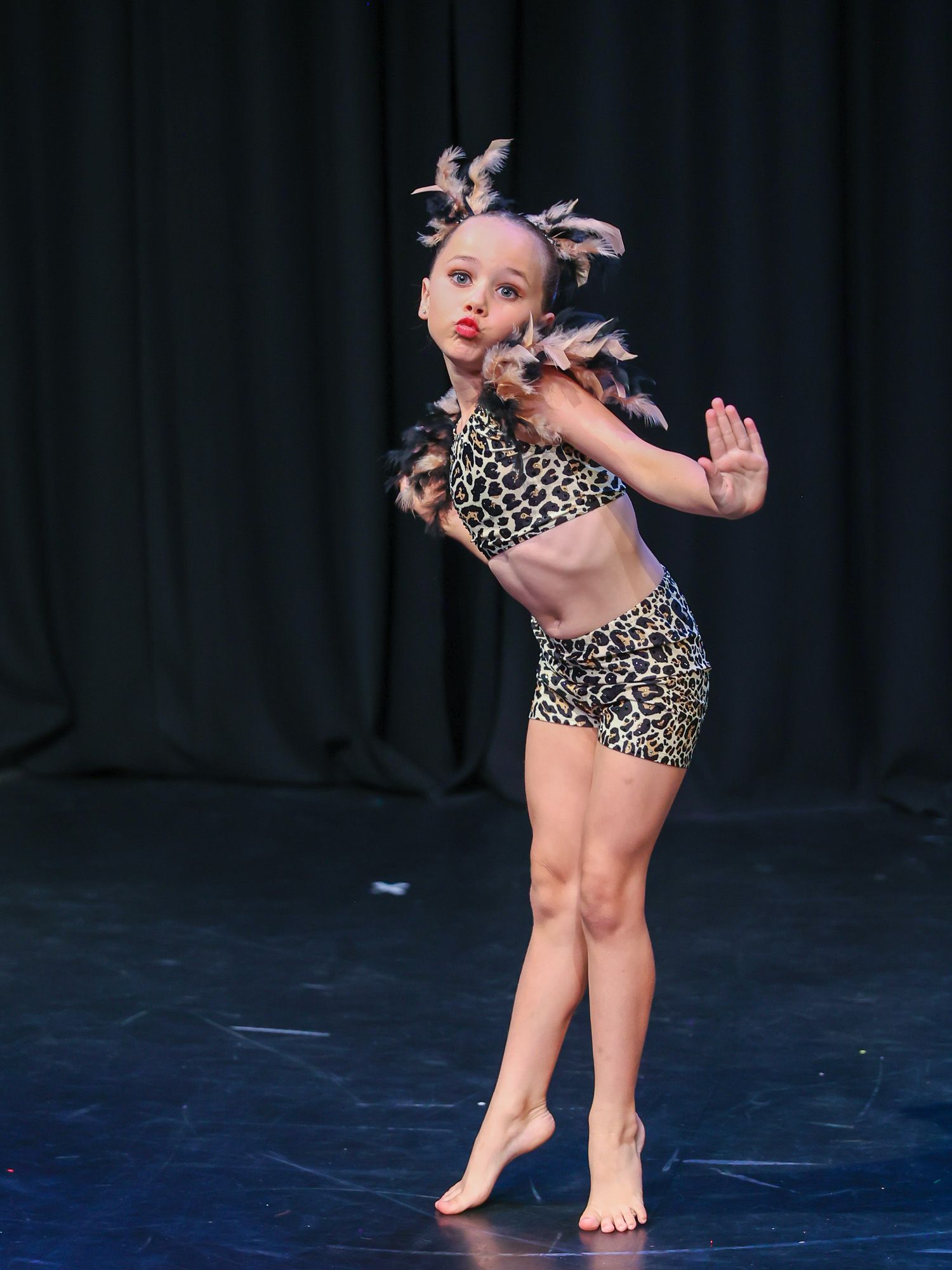 Young girl performing on stage in leopard print costume with feather accessories, striking a dramatic dance pose.