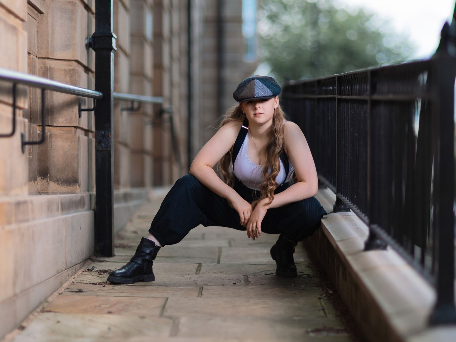 Young woman in flat cap and black outfit crouching on cobblestone path beside historic stone building.
