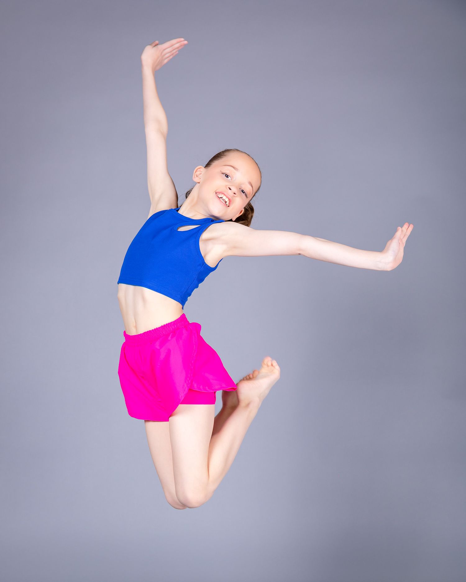 Dancer in blue crop top and pink shorts leaps gracefully through the air against a gray studio backdrop.