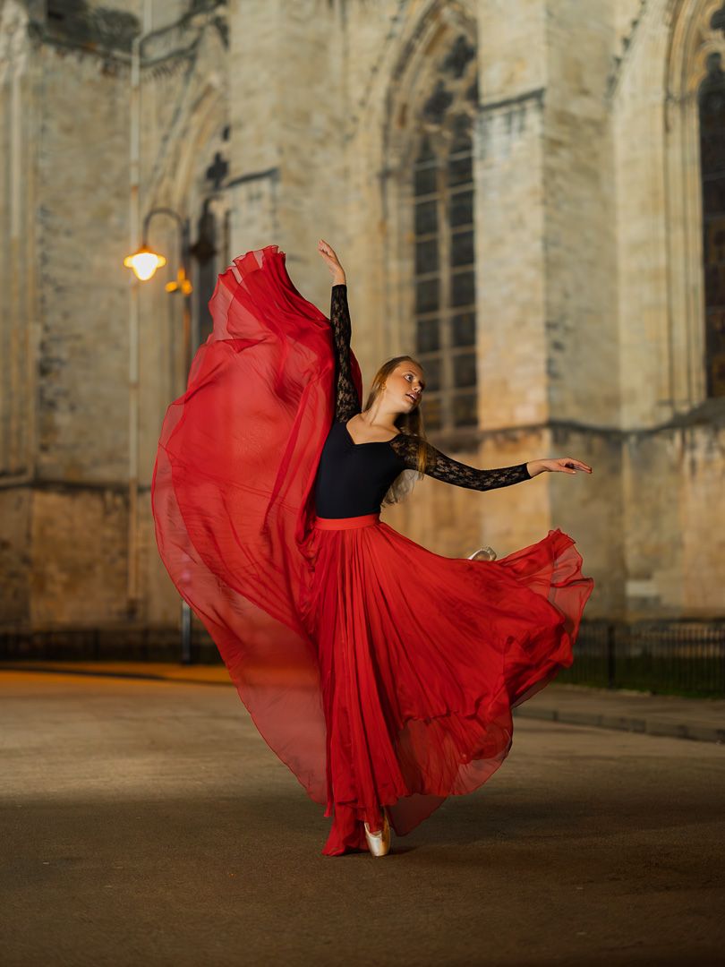 A ballet dancer in a flowing red skirt and black lace top performs gracefully outdoors near a historic stone building.